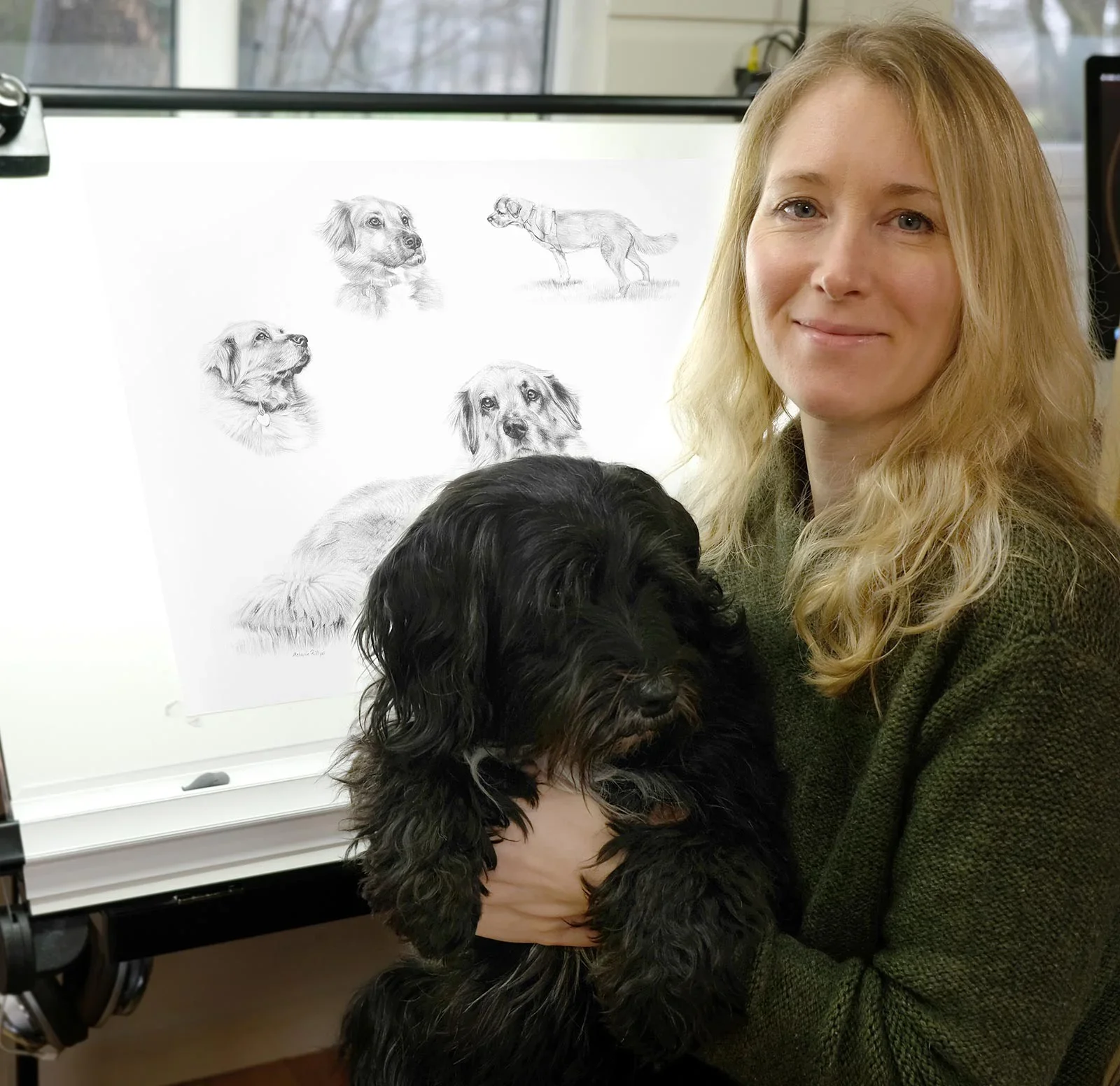 Life Story Pet Portraits in Pencil by Melanie Phillips at her easel with a portrait and dog
